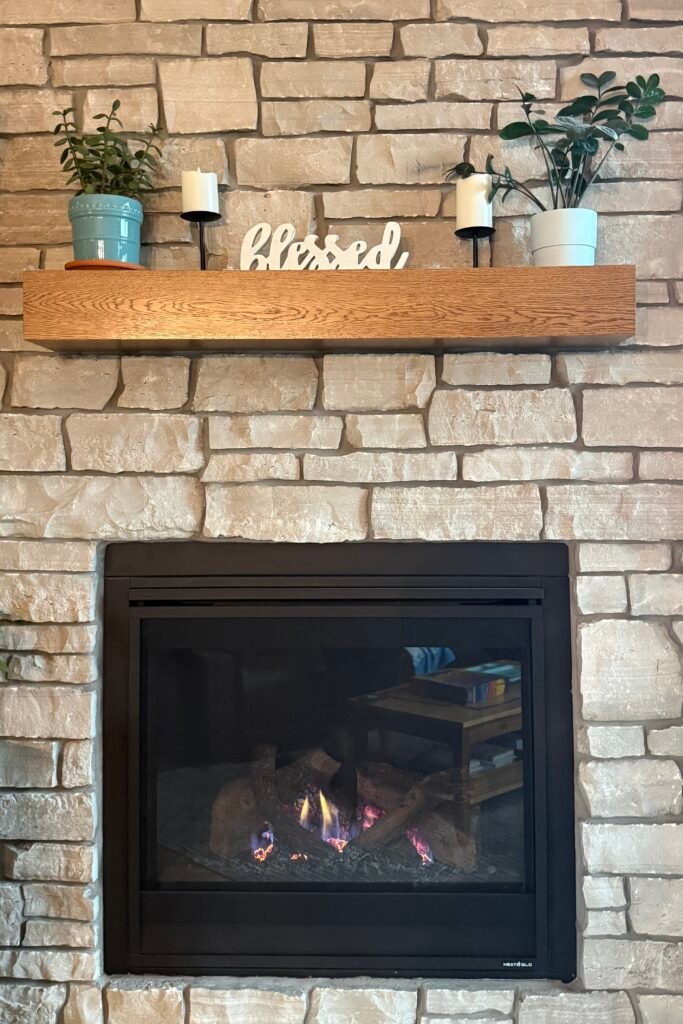 A portrait view of a light stacked-stone fireplace hearth with a lit fire, a natural oak mantel decorated with green plants, and a wooden "blessed" sign.