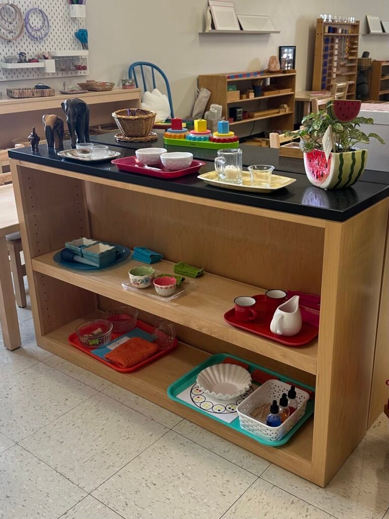 An open child-sized shelving unit filled with organized activity trays in a bright Montessori learning environment.