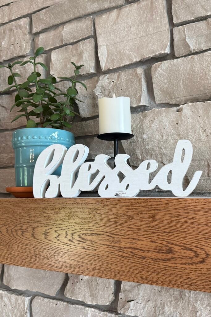 White decorative “Blessed” sign displayed on a wooden mantel against a stone fireplace, with a teal potted plant and white candle.