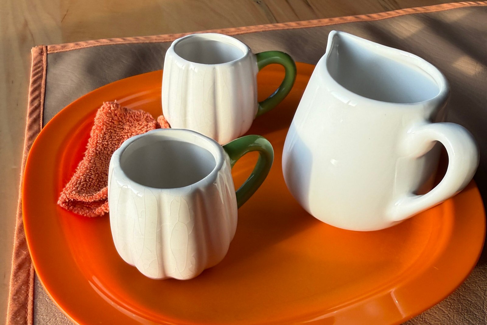 A Montessori practical life tray with a white ceramic pitcher, two small handled cups, and a folded cloth arranged on a bright orange tray atop a neutral work mat on a wooden table in warm natural sunlight.